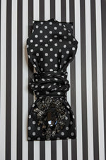 Load image into Gallery viewer, Black polka dot necktie necklace with a decorative clasp on a black and white striped background
