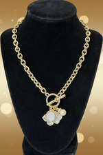 Load image into Gallery viewer, A gold chain necklace with a toggle clasp and a flower-shaped charm paved with clear stones and accented with a small pearl, displayed on a black velvet jewelry bust 
