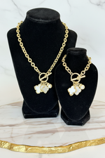 Load image into Gallery viewer, A gold-tone chain necklaces with matching bracelet displayed on black velvet jewelry busts. Each piece features a toggle clasp and dangling charms including a white iridescent flower, a small white pearl, and a clear rhinestone cluster.

