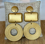 Load image into Gallery viewer, A pair of chunky gold-toned geometric dangle earrings displayed on a woven beige fabric stand over a wooden surface. Each earring has a round disc, a rectangular bar, and a large concentric-ring circle drop.
