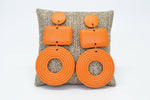Load image into Gallery viewer, A pair of chunky orange geometric drop earrings displayed on a woven cushion. Each earring features a small round top, a rectangular middle section, and a large circular bottom piece with concentric ring detailing.
