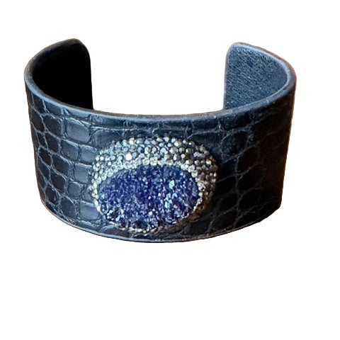 Black crocodile-patterned cuff bracelet with a blue gemstone on a wooden surface