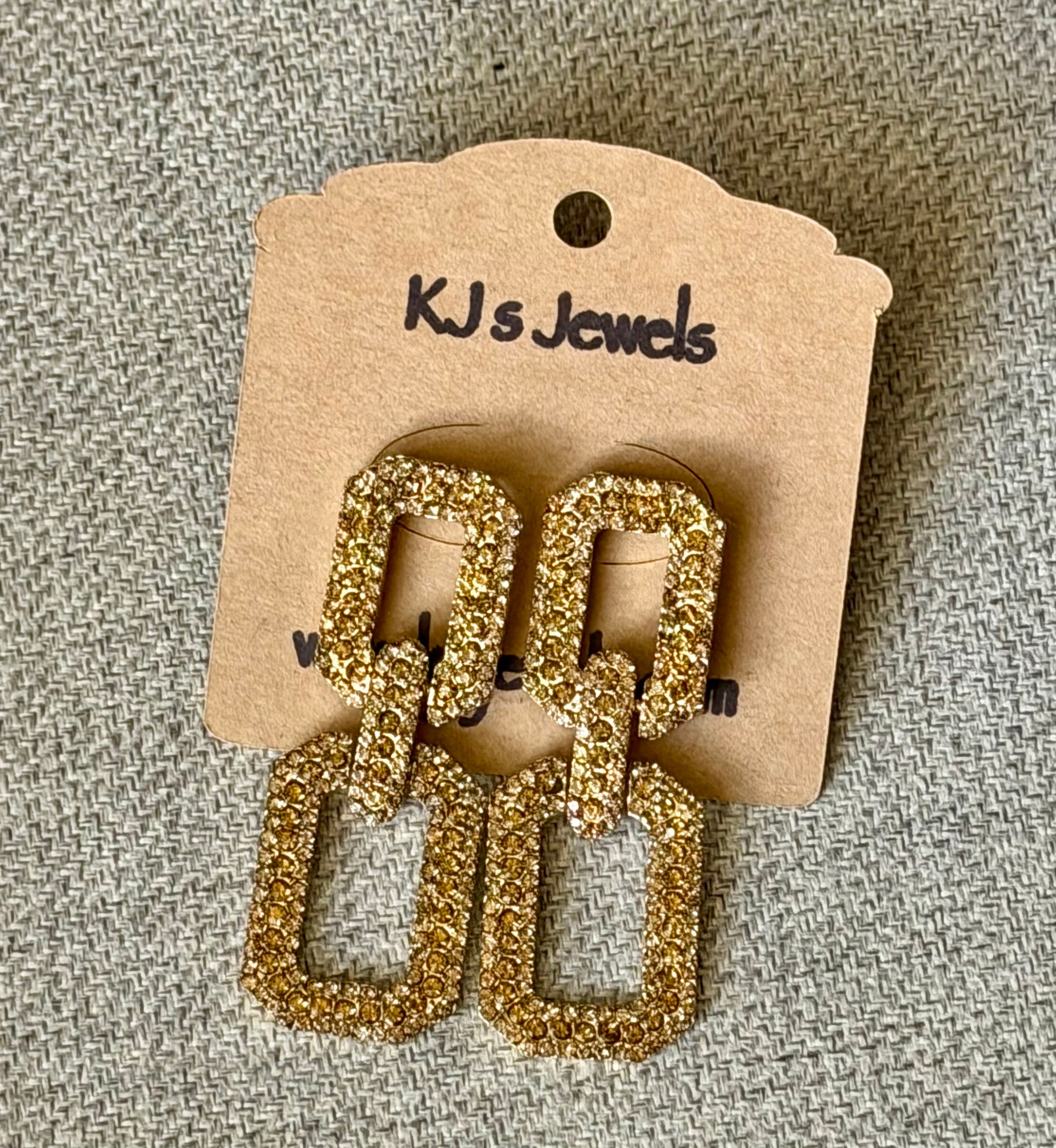 Gold glittery earrings on a KJ's Jewels branded card against a textured gray background