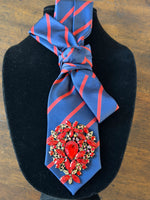 Load image into Gallery viewer, Blue and red striped tie with a decorative brooch on a black stand.
