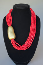 Load image into Gallery viewer, A multi-strand red beaded statement necklace with a large polished gold pendant, displayed on a black velvet jewelry bust.
