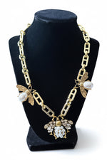 Load image into Gallery viewer, Gold-tone statement necklace displayed on a black velvet jewelry bust, featuring a chunky link chain, bee motifs with white pearls, and a central bee embellished with rhinestones and white faux pearls.
